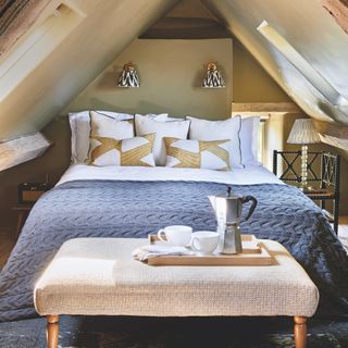 attic bedroom in Cotswolds cottage with wall lights above bed, skylights in roof, upholstered coffee table at base of bed