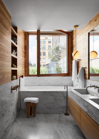 a bathroom with marble used on the flooring, bath tub and counters. There are wooden elements used in the bathroom too. A large window is open and draws in natural lighting.