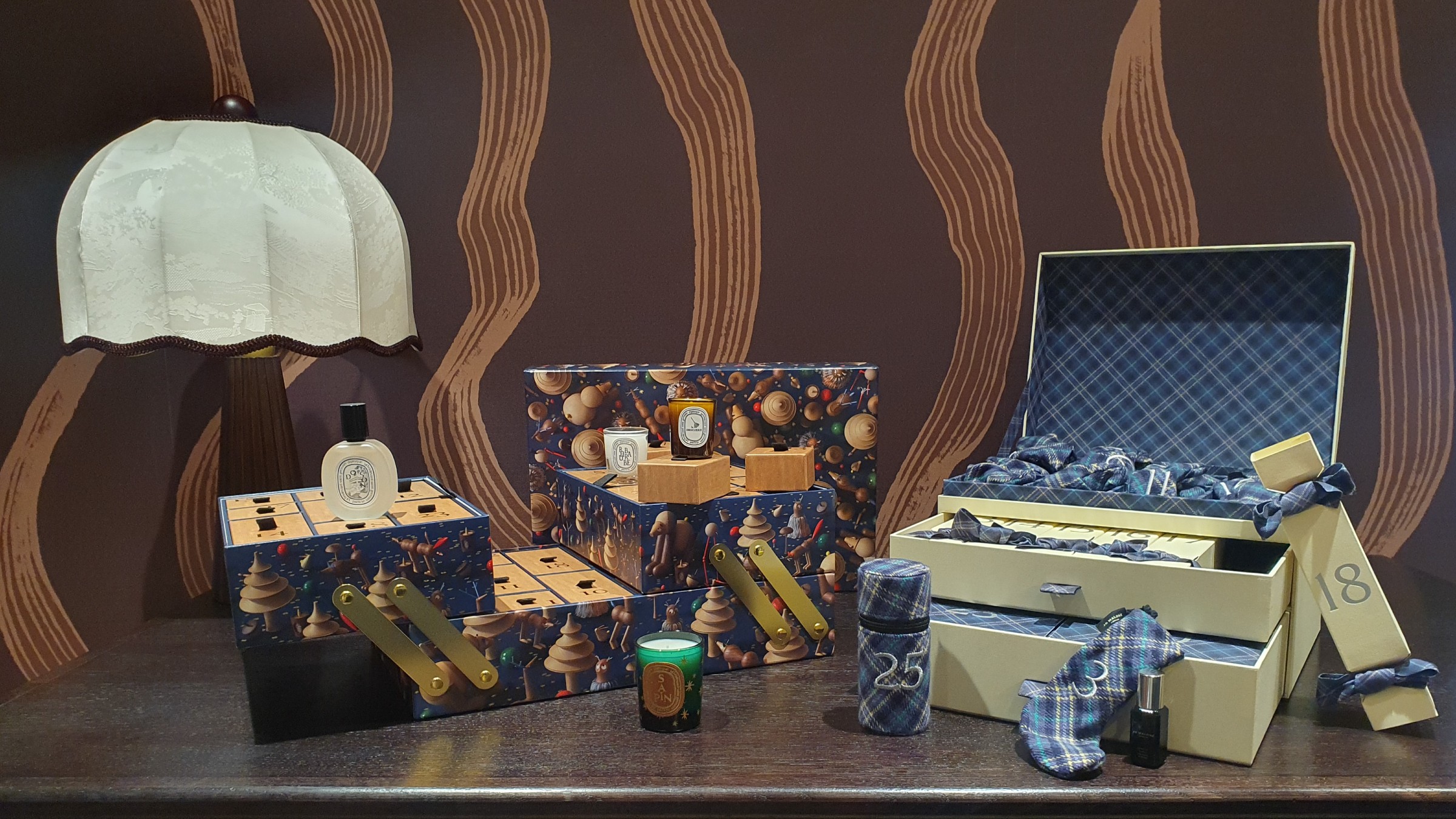 An open Diptyque and Jo Malone luxury advent calendar for 2026 with a few products taken out set on a dark wood table with a brown patterned wallpaper in the background