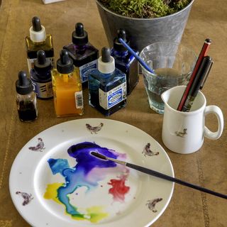 Plate on wooden counter with various colours of paint on and paint brush next to glass containers with different colors of paint in