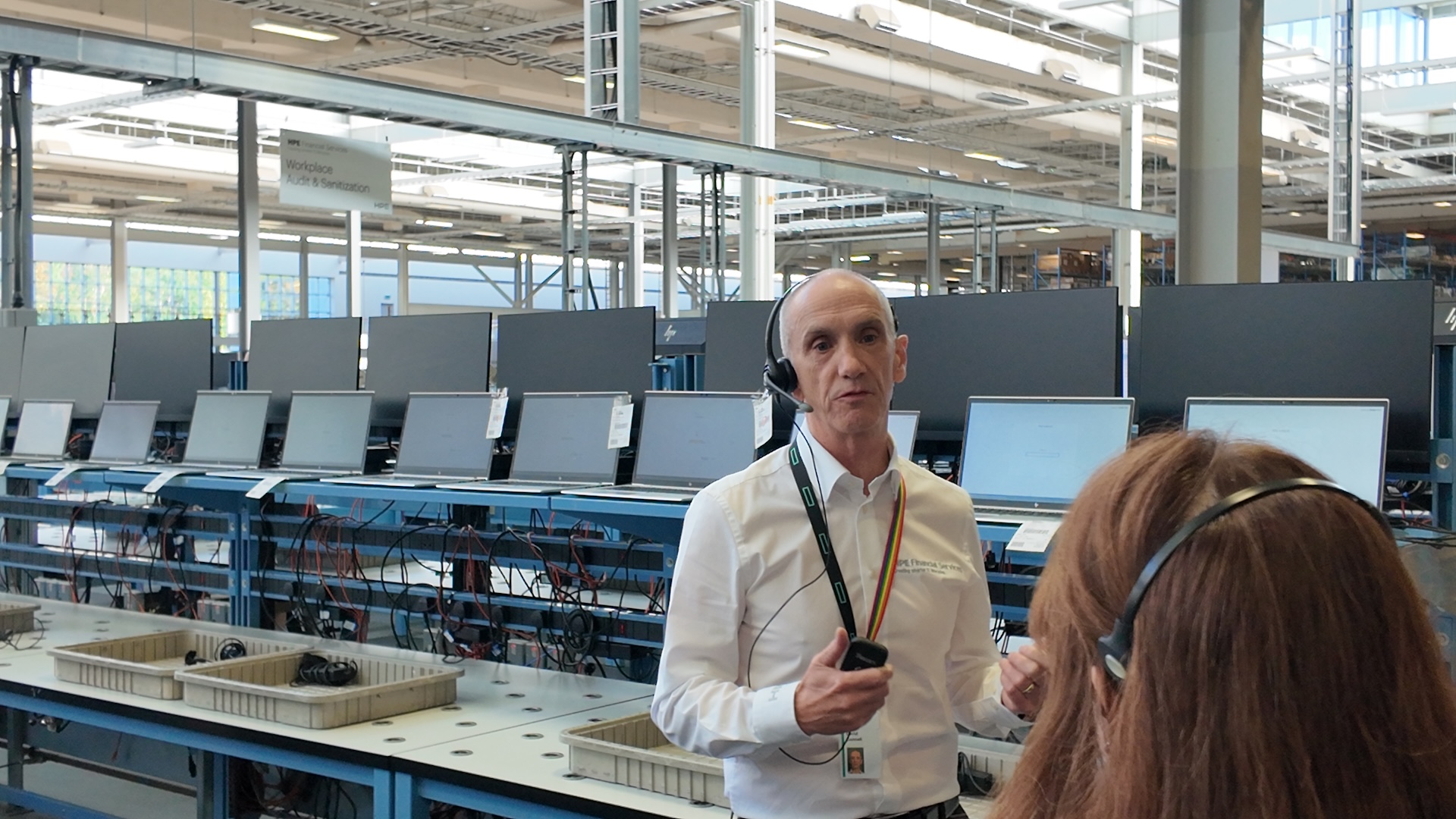 A man in a white shirt and headset speaks to a tour group with netbooks set up in a neat row behind him for sanitization.