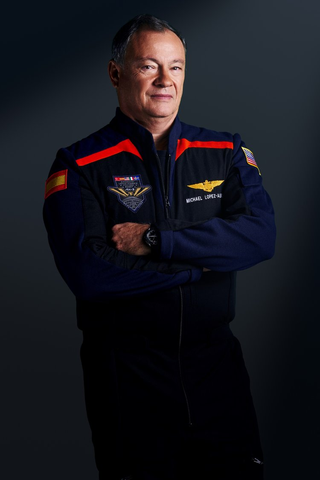 A man stands with arms crossed, wearing dark blue jumpsuits with red accented shoulder wings. He looks serious.