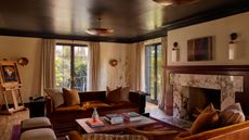 Living room with a brown-painted ceiling, cream walls, a large marble fireplace, two burnt-orange velvet sofas, a wooden coffee table, and an easel holding an oil painting in the corner.