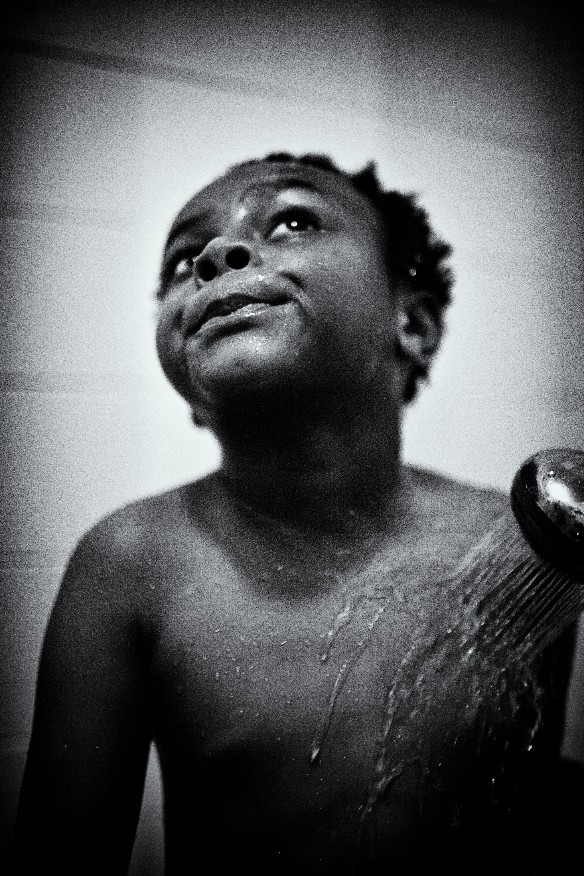 Uma foto em close em preto e branco de um menino sorrindo e olhando para cima enquanto a água de um chuveiro enxagua seu ombro.