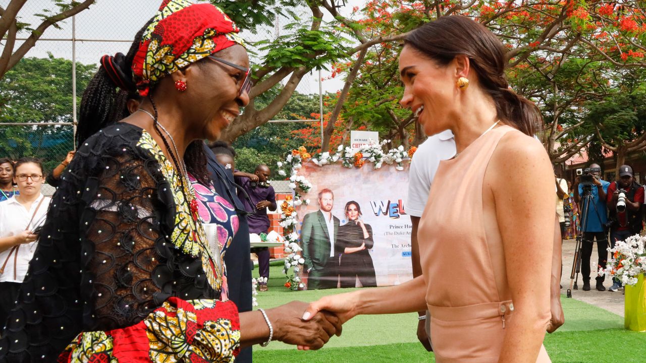 Meghan Markle shakes hands with a woman in Nigeria while wearing a peach sundress by heidi merrick