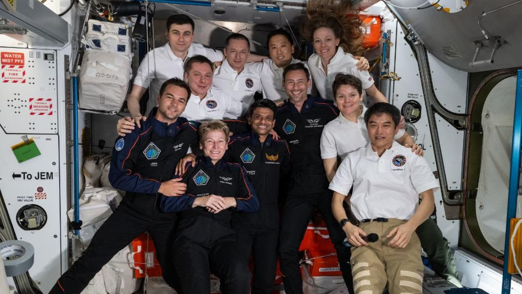 photo of 11 smiling people in a crowded space module. seven are wearing white polos and the other four are wearing dark blue flight suits