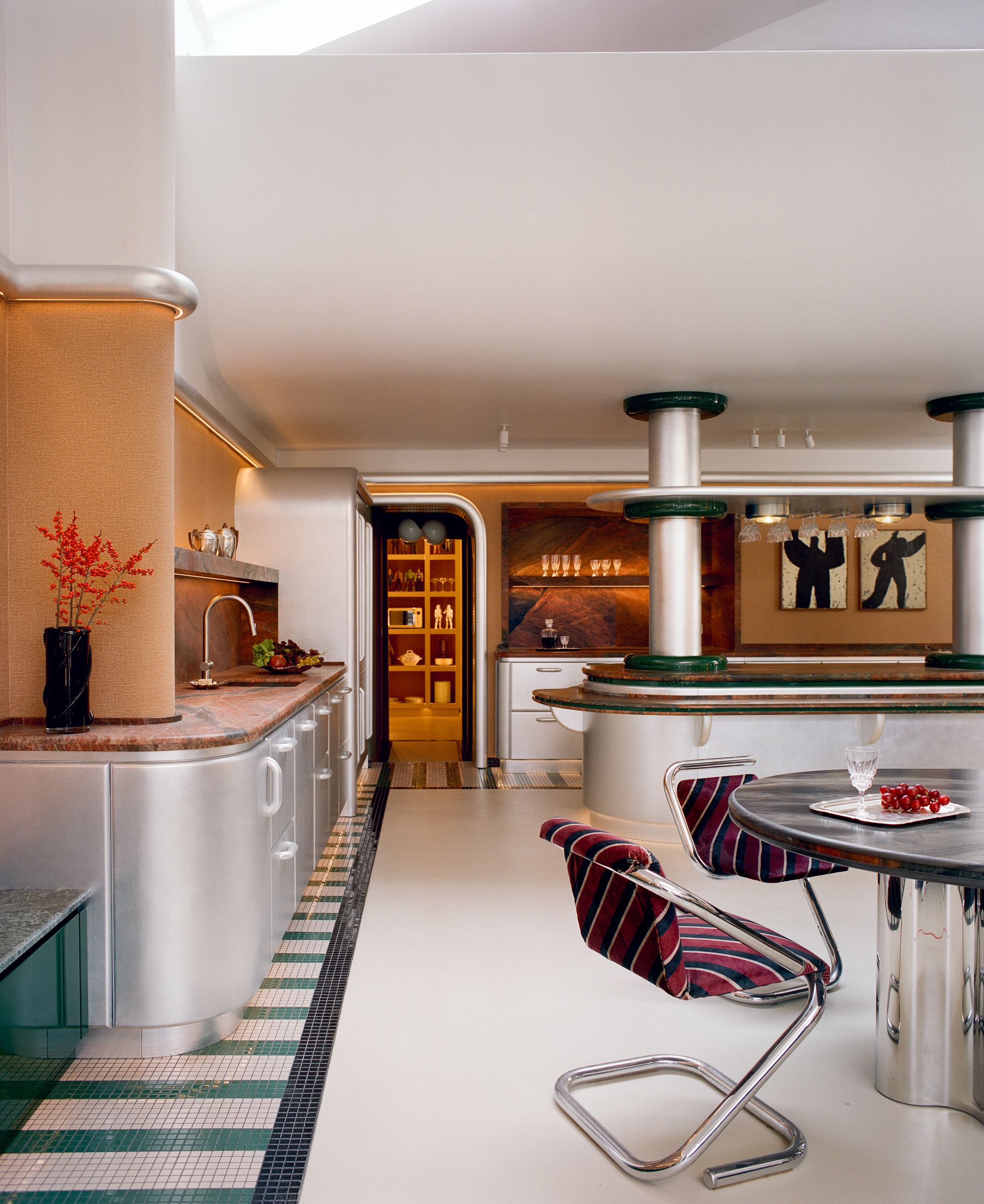 Image of a large Space Age-inspired kitchen with chrome cabinetry and green mosaic tile floors. The walls are orange and white color block, and there is orange-ish red marble on the counters.