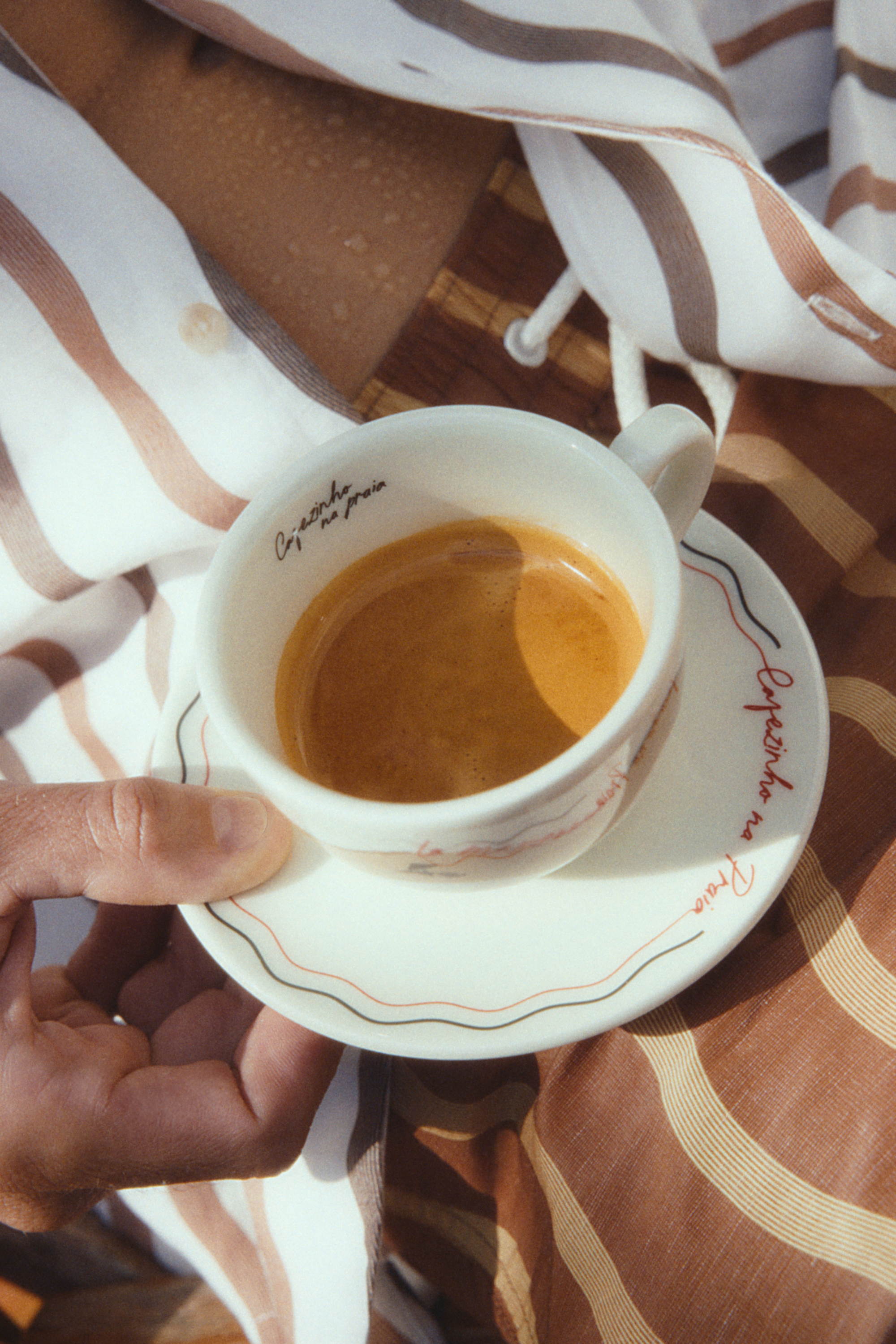 Image of a hand holding a white espresso cup and saucer with espresso in it over a brown and white striped shirt and bathing suit.