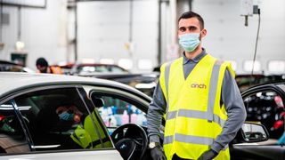 Man in a mask and high-visibility vest standing in front of a car in a warehouse