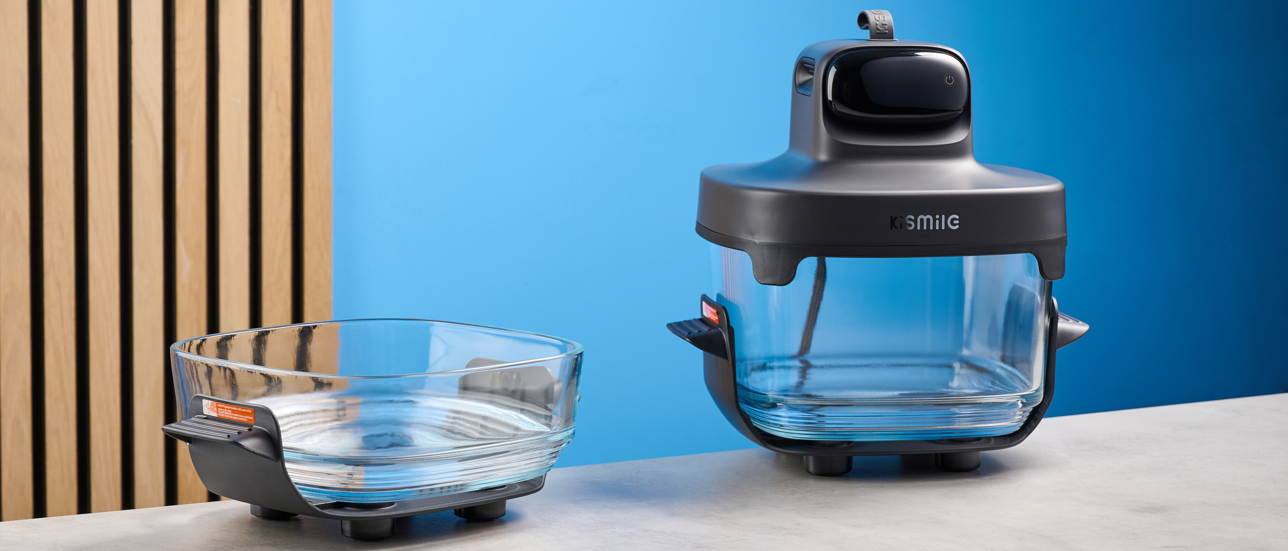 the kismile roasti air fryer with a glass cooking container photographed against a blue and wood background, showing the heating element lid