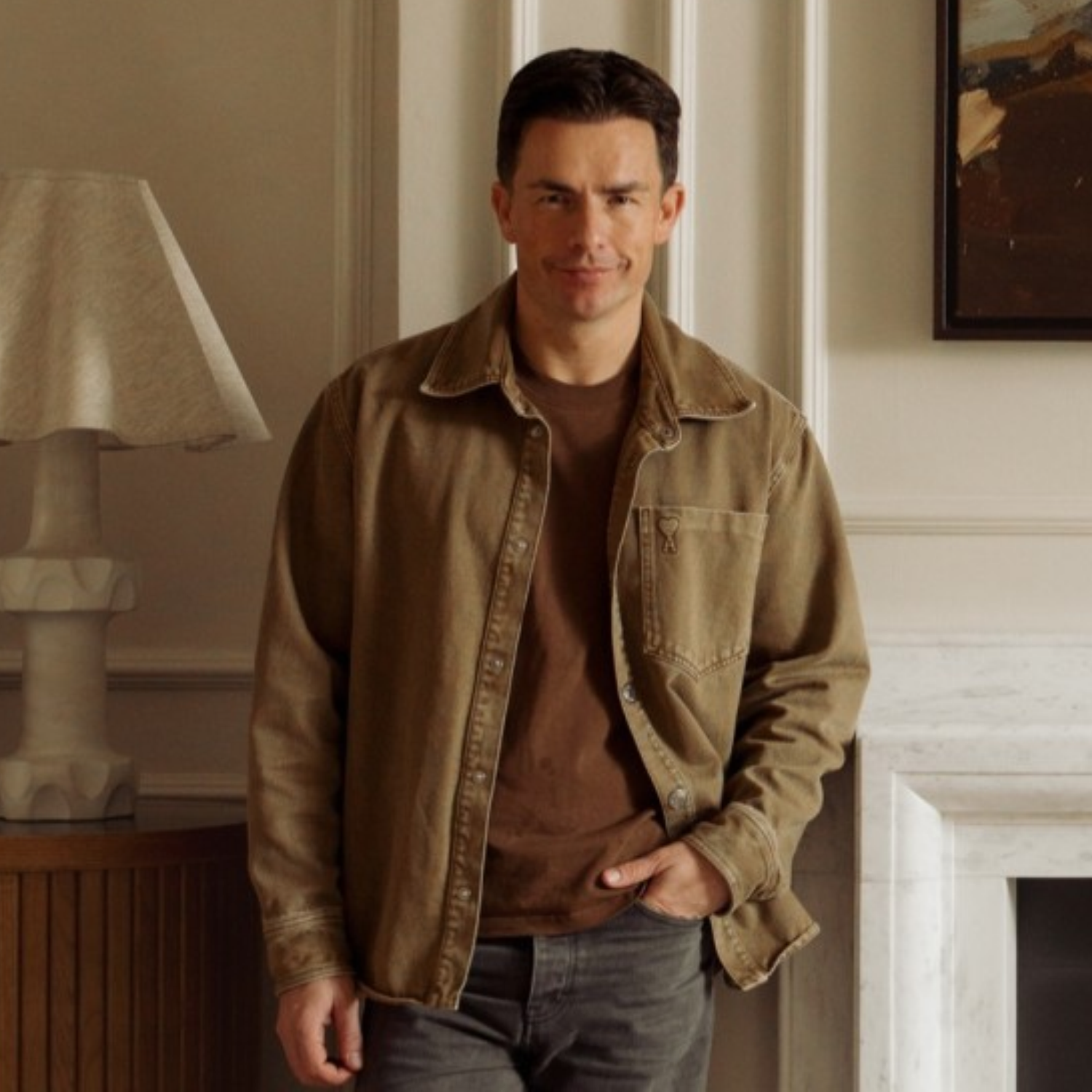 A man in a brown shirt layered with a t-shirt, leaning against a mantel