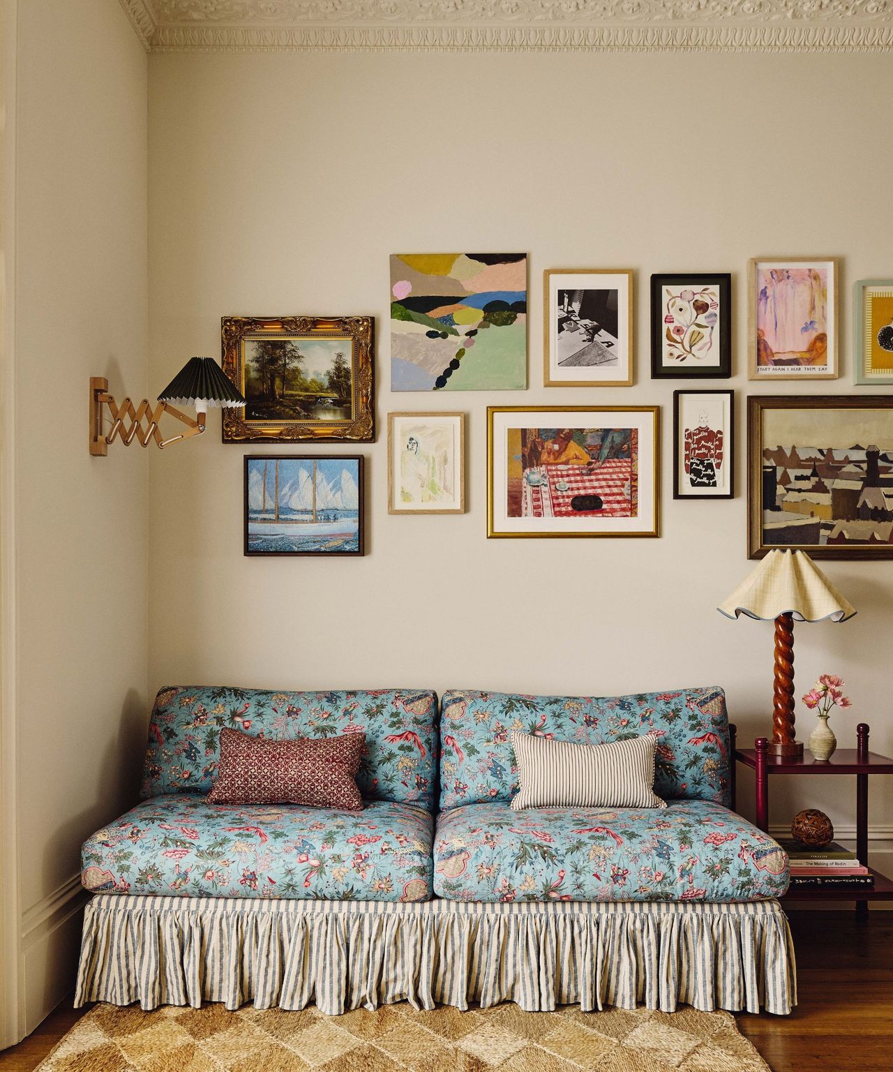 A sitting area with cream walls and vintage-style floral sofa with a striped skirt. Above it, a gallery wall displays a mix of colorful contemporary and classic framed artworks in various sizes.