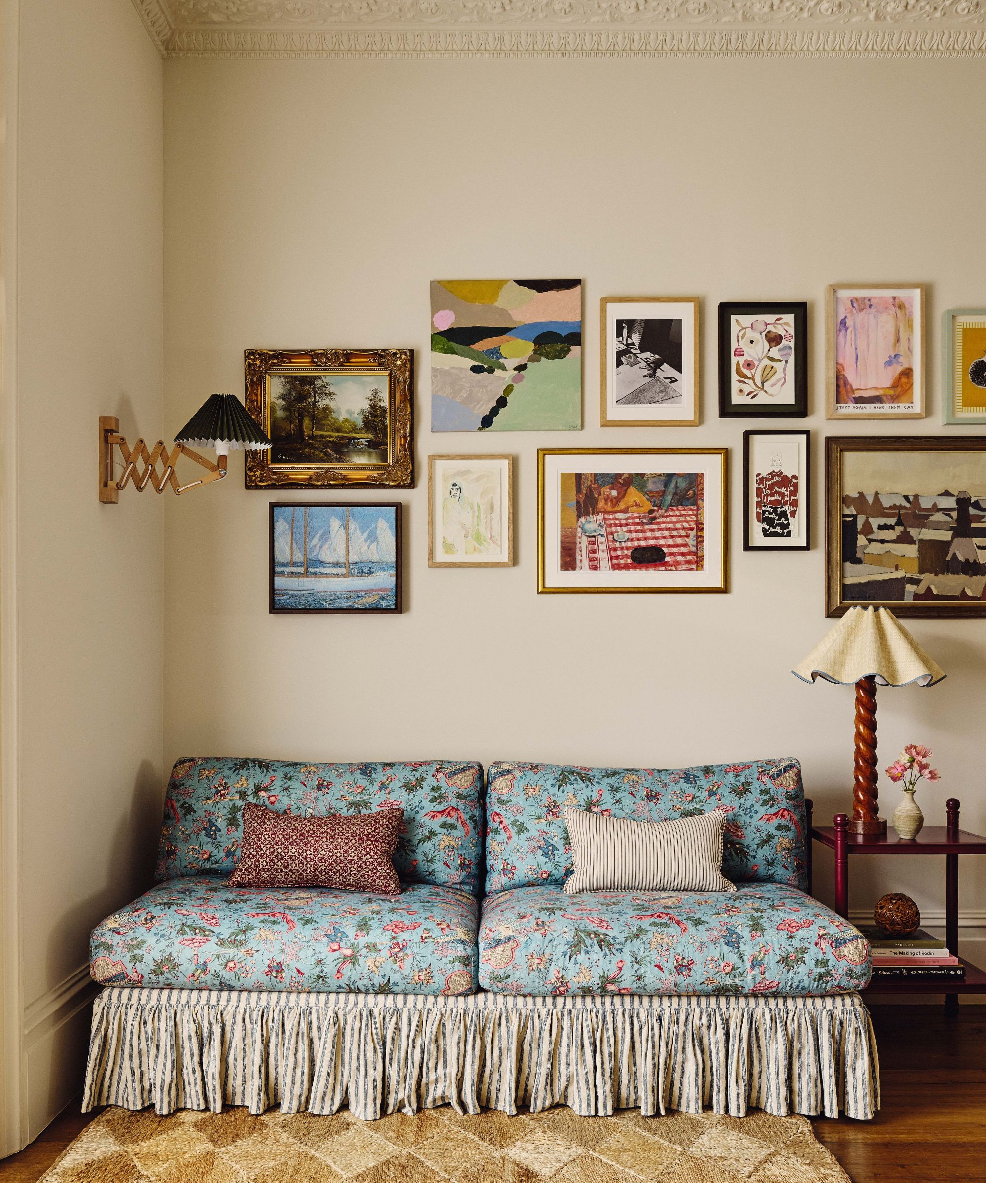 A sitting area with cream walls and vintage-style floral sofa with a striped skirt. Above it, a gallery wall displays a mix of colorful contemporary and classic framed artworks in various sizes.