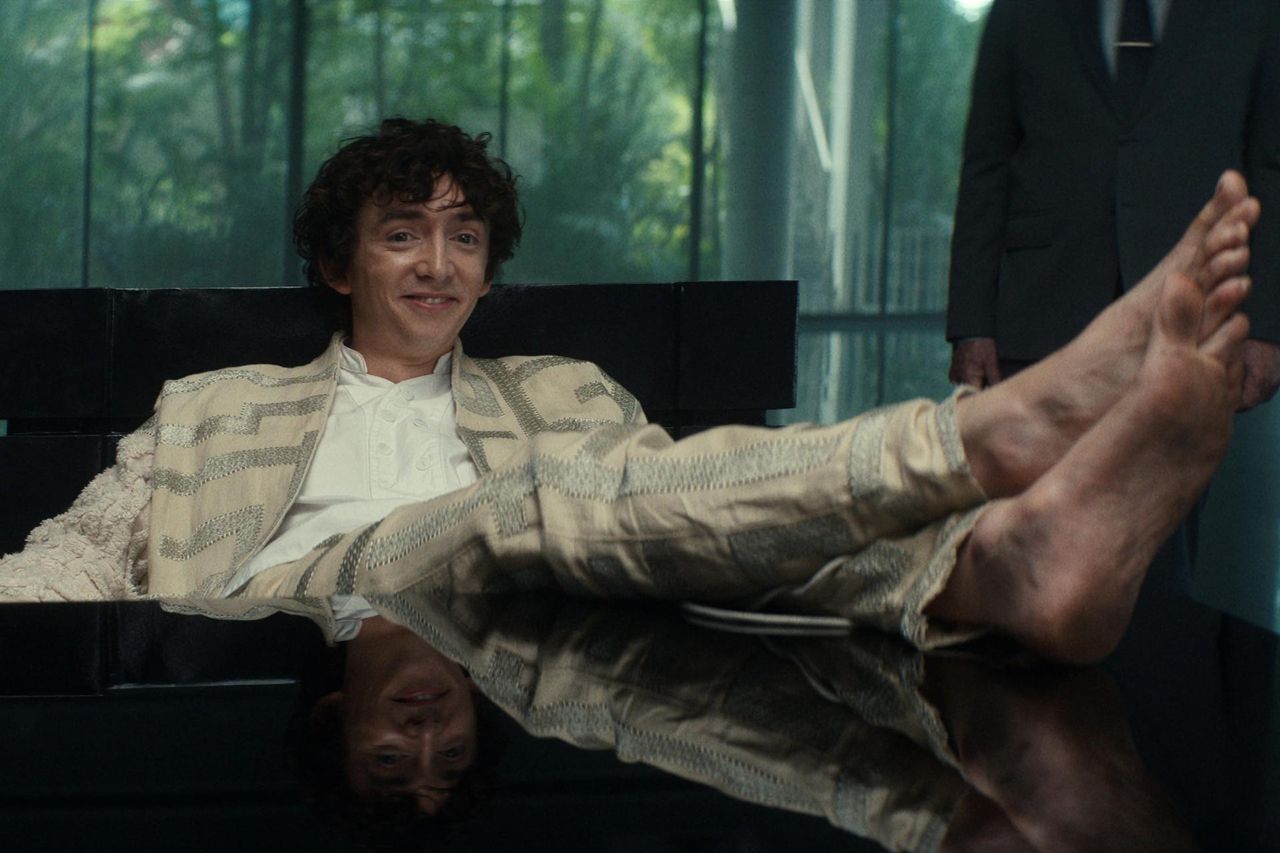 Still from the TV show Alien: Earth. A grinning man (wearing a beige and silver suit jacket and pants) is sitting at a desk with his bare feet on the desk.