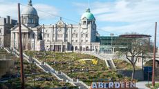 A wide shot of Union Terrace Gardens in Aberdeen, with people walking down the steps and a large official building in the background. In the lower foreground, a large sign reading "ABERDEEN" can be seen, with people milling about it. Decorative: the scene is filled with greenery and the sky is blue and sunny.