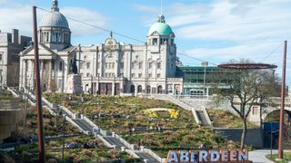 A wide shot of Union Terrace Gardens in Aberdeen, with people walking down the steps and a large official building in the background. In the lower foreground, a large sign reading "ABERDEEN" can be seen, with people milling about it. Decorative: the scene is filled with greenery and the sky is blue and sunny.