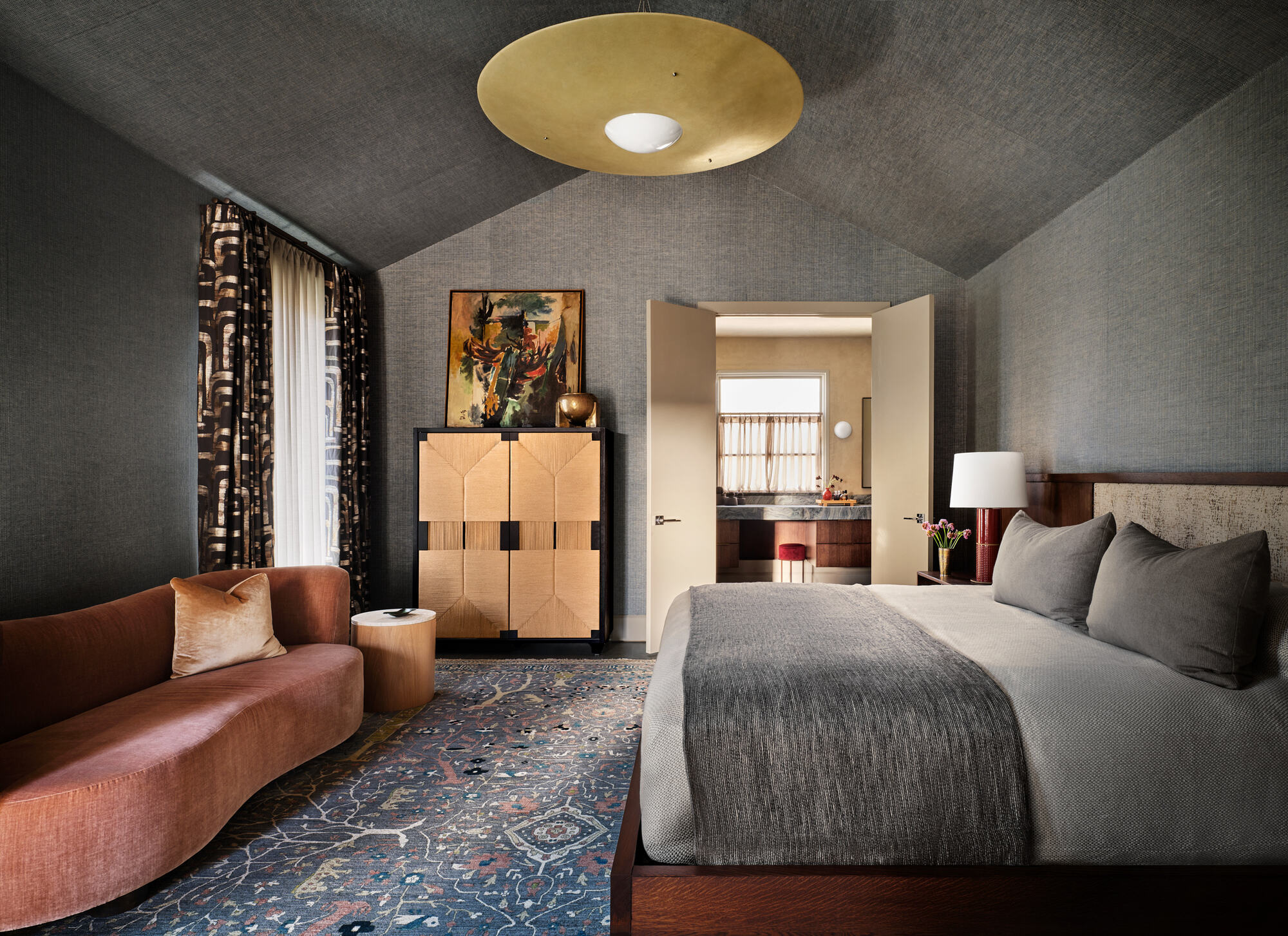 modern bedroom with gray wallcovering which continues across the roof, a gold pendant light, a rust-colored sofa, patterned blue rug, gray bedding, freestanding cabinet with artwork leaning above, and doors through to the ensuite