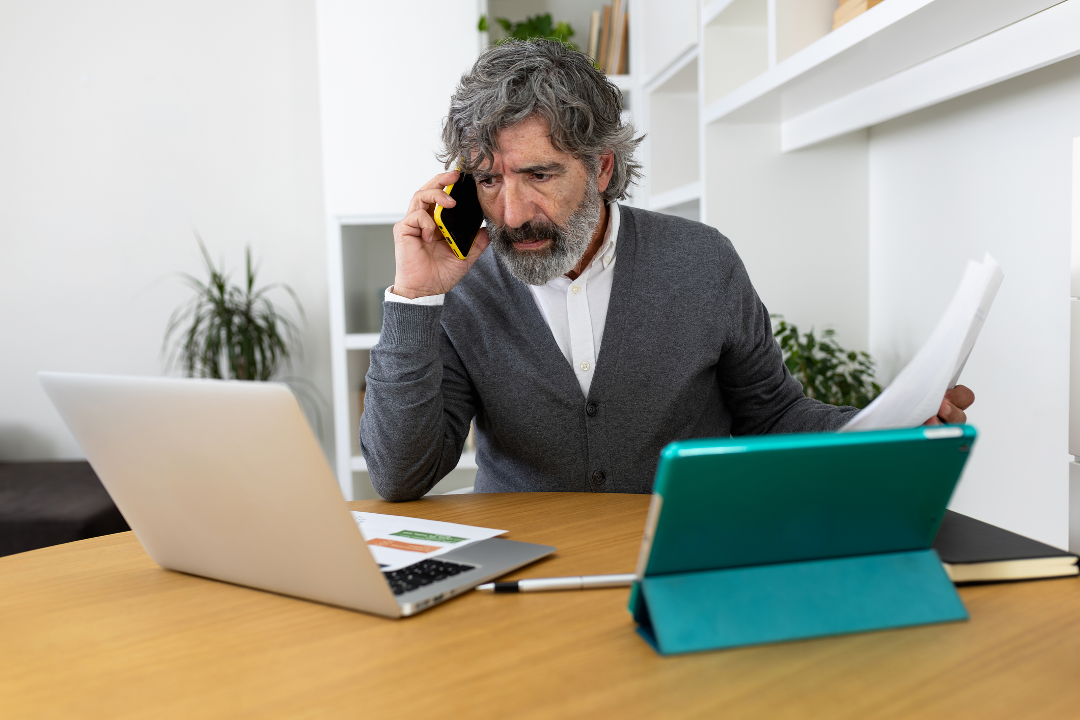 an older man is on the phone while working on his finances with two laptops and charts on his desk