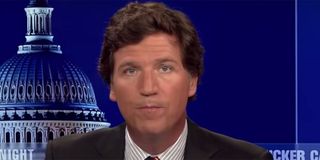 Tucker Carlson with a suit on sitting at his desk and talking to viewers.