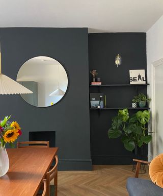 A living room with a white ceiling, a black accent wall with a mirror with shelves on it and a wooden dining table in front