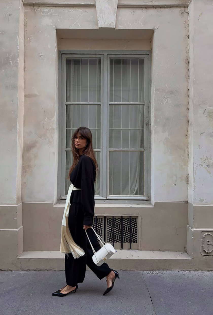 A woman wearing a black crewneck sweater with a white silk scarf tied around the waist, styled with black balloon pants, black pumps, and a white bag.