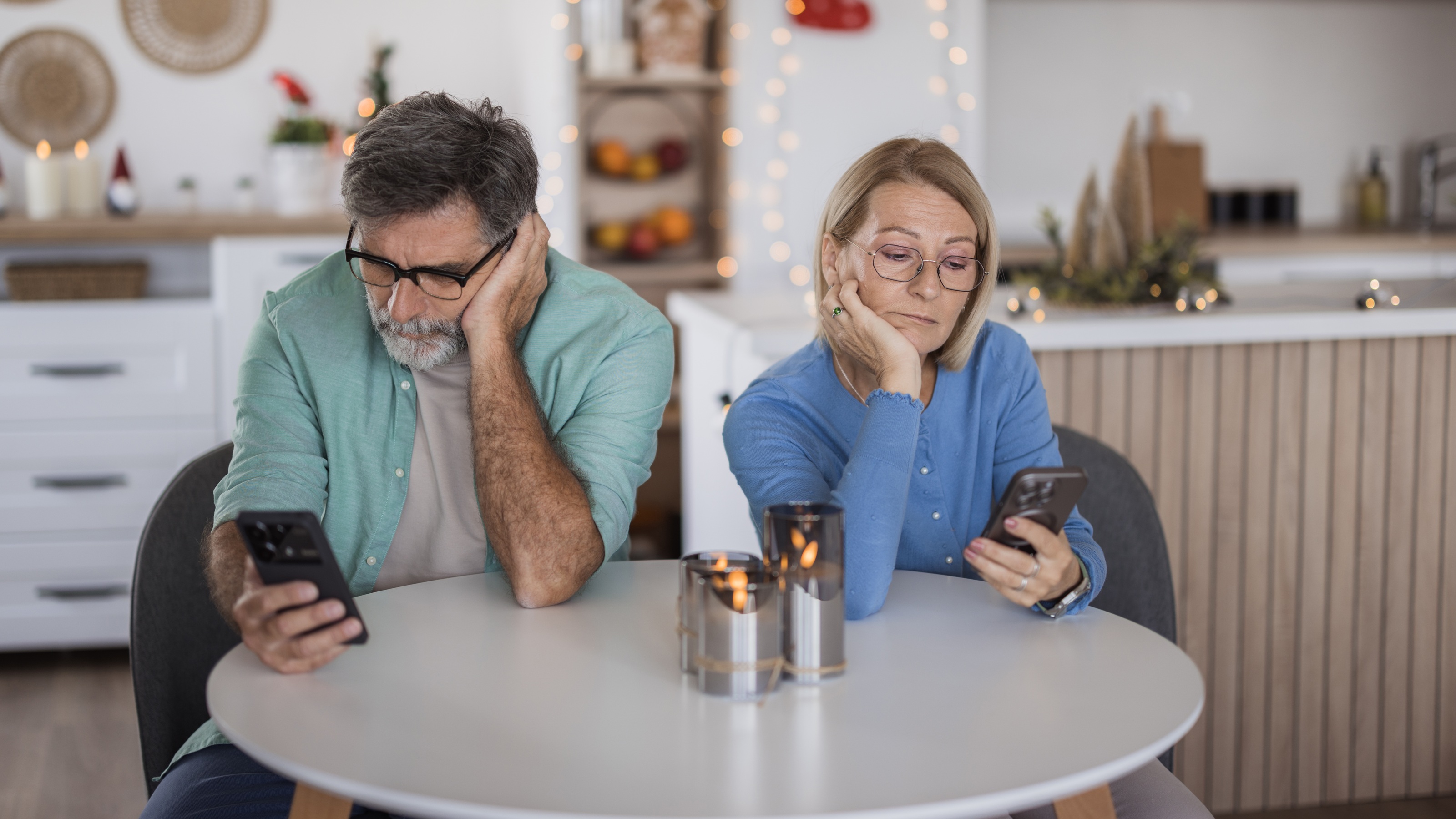 An older couple look bored as they sit at their kitchen table looking at their phones.