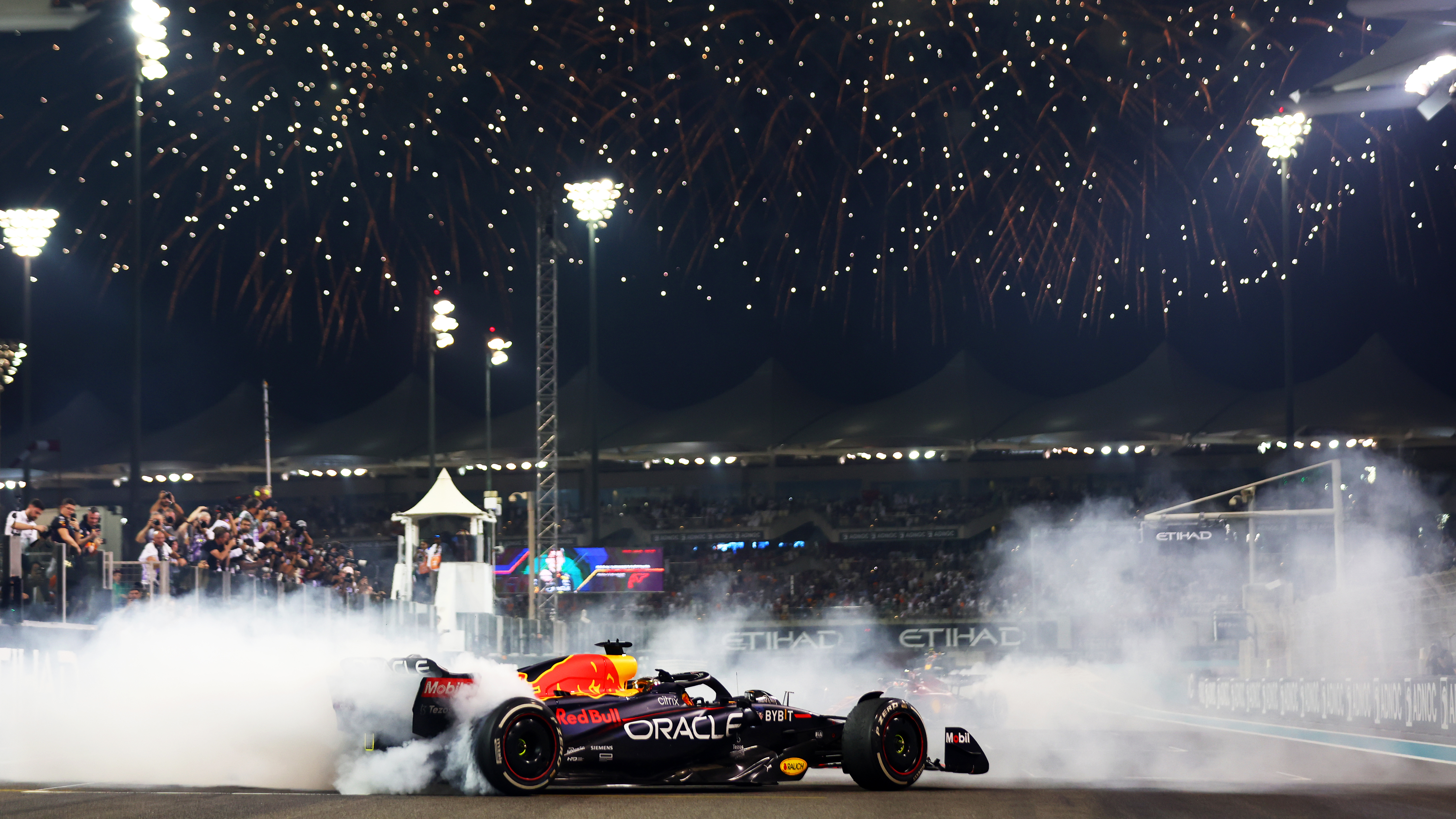 An F1 car doing a victory donut on a track, surrounded by tyre smoke. Fireworks are going off in the night sky above the track. Decorative: the car bears the logos for Red Bull and Oracle, as it is the car of Oracle Red Bull Driver Max Verstappen.