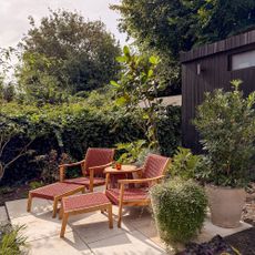 Two wooden and read woven recliner chairs with some table on patio area by hedge in garden