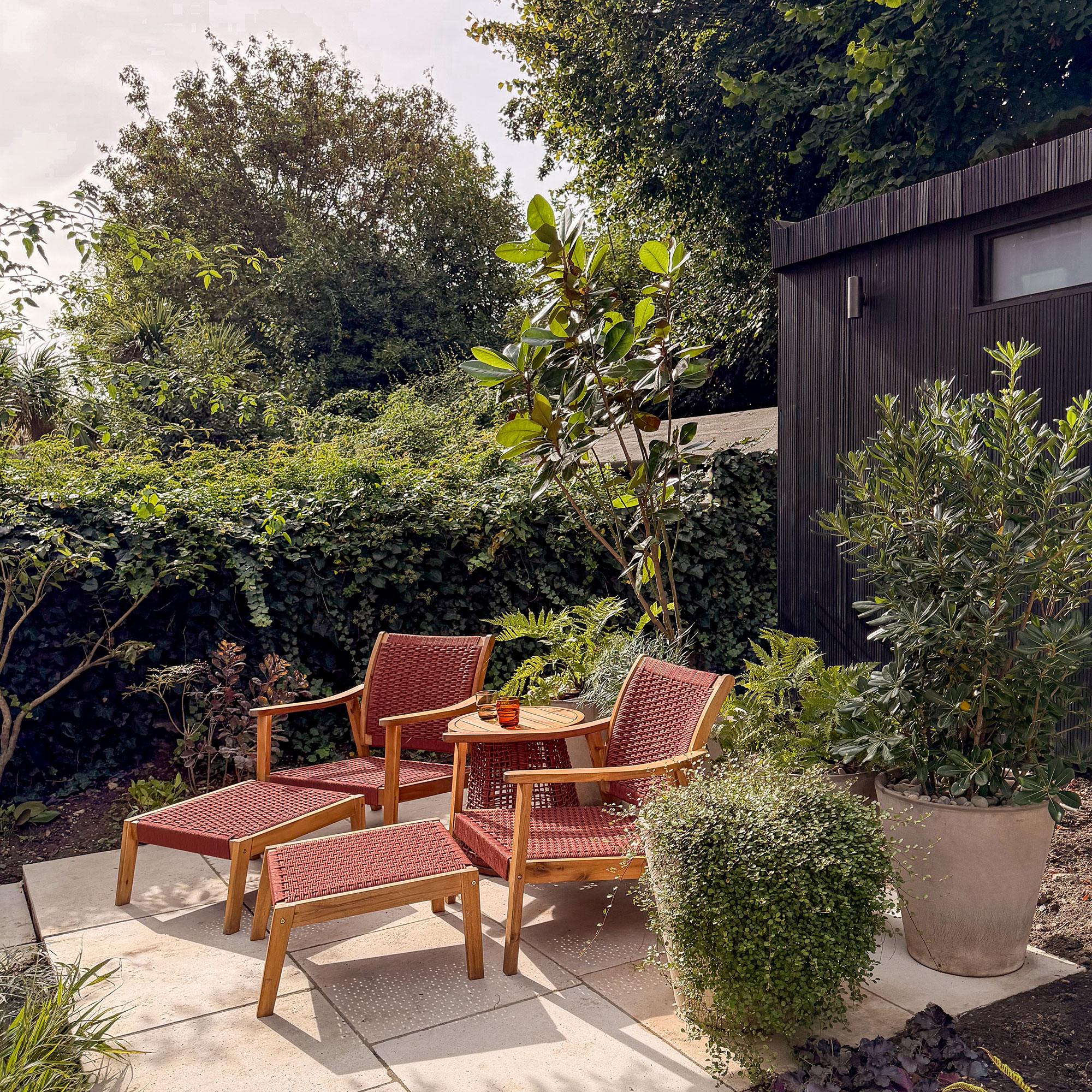 Two wooden and read woven recliner chairs with some table on patio area by hedge in garden