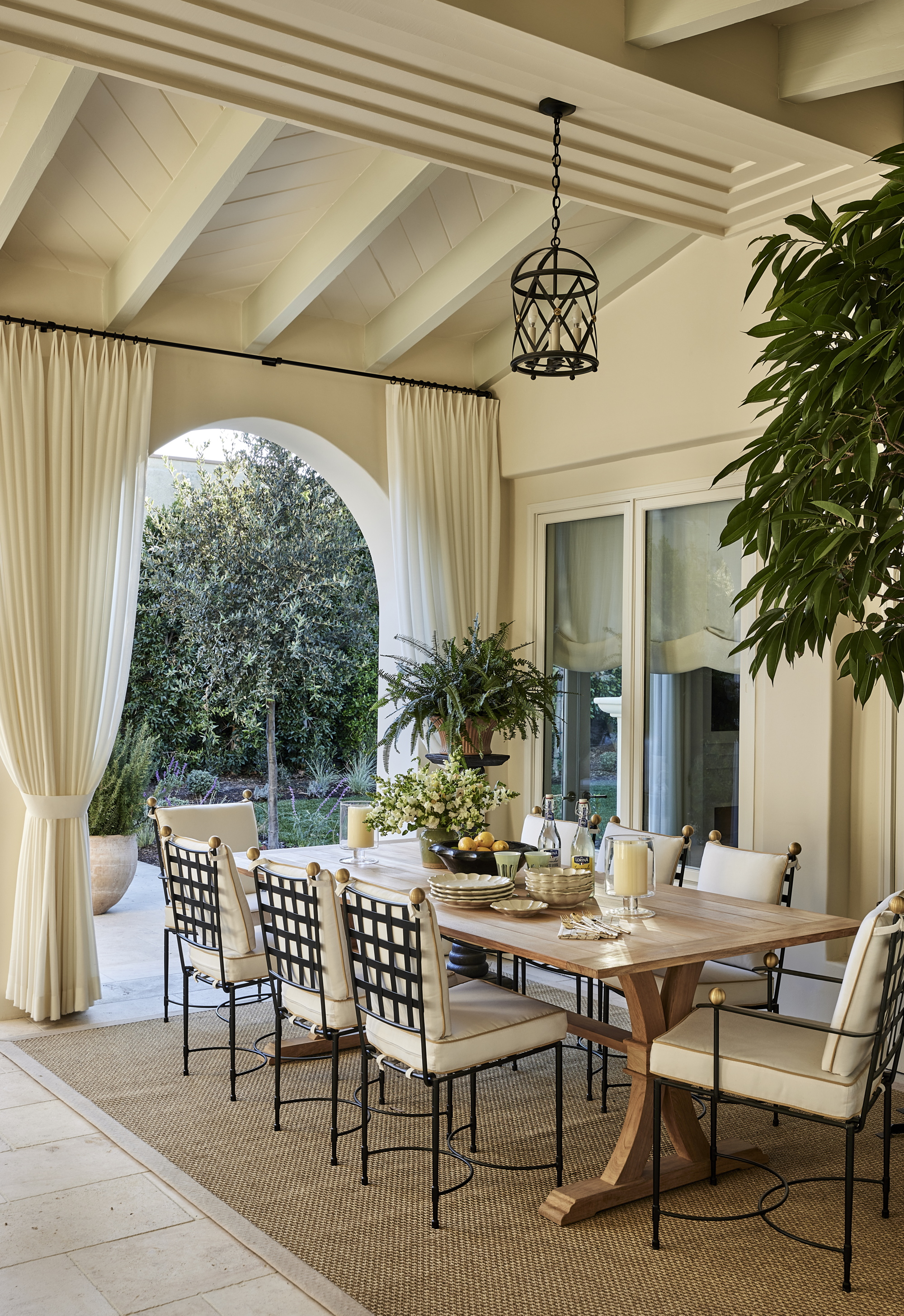 A sheltered outdoor patio area with a large dining table, chairs, and sheer curtains hanging above an arch