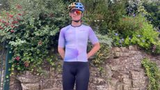 The purple Unisex Santini Ombra cycling jersey seen on a merged image of half male and half female rider with a stone wall and planting behind them 