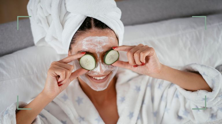 Woman holding cucumber slices to her puffy eyes and smiling wearing a facemask and a dressing gown