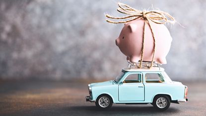 A piggy bank is tied to the top of an older model (toy) car.