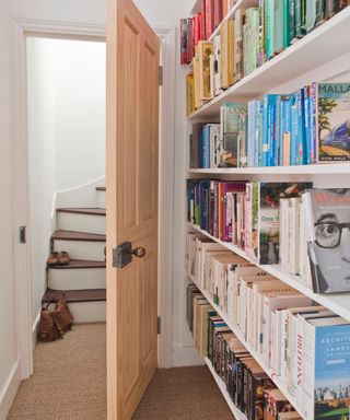 Hallway and landing with narrow curving staircase leading up to loft, modern pine door with antique door handle, large white wood open book shelves, brown hessian carpet,