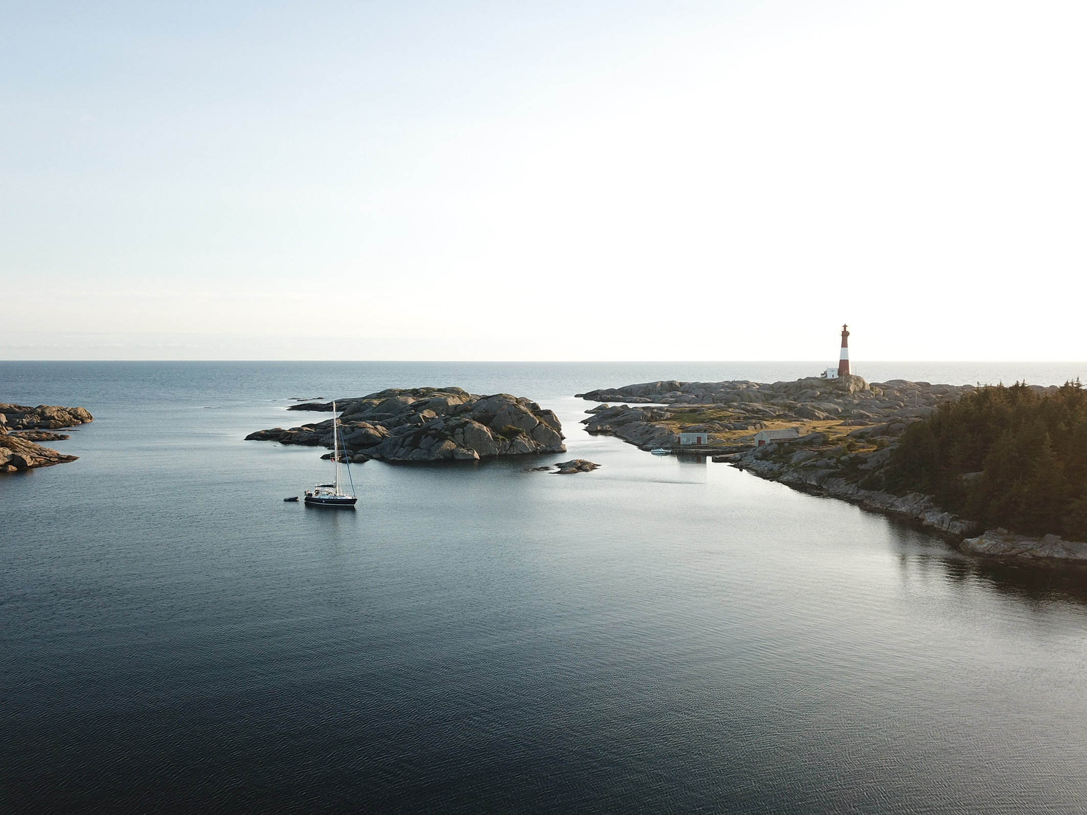 A sailboat anchored in calm waters near rocky shores and a tall lighthouse against a serene sky