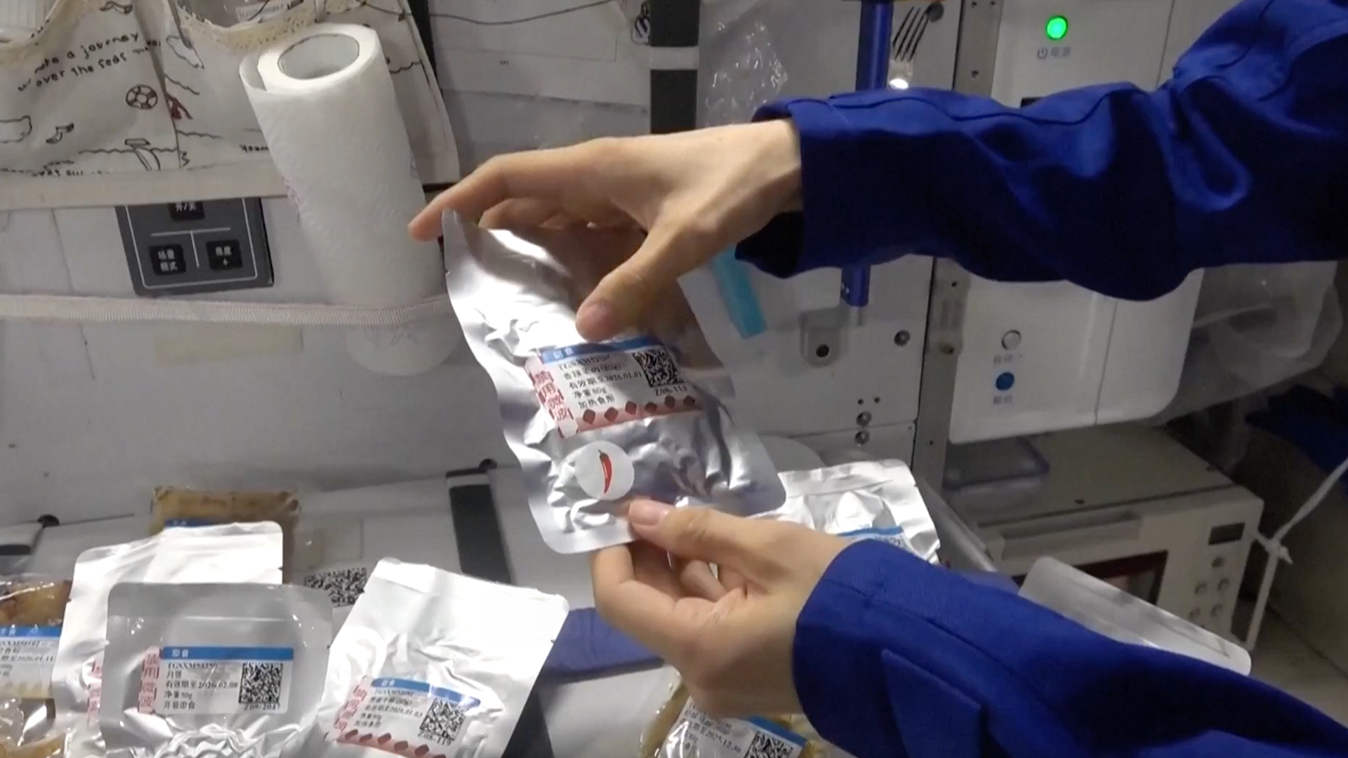 people in blue flight suits open foil-wrapped packages of food in a cramped rectangular laboratory
