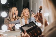 Two young woman sit in front of an illuminated mirror and apply make-up