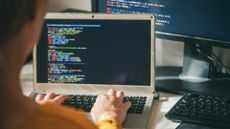 Software developer using AI coding tools on a laptop computer with source code pictured on screen and desktop monitor in background.