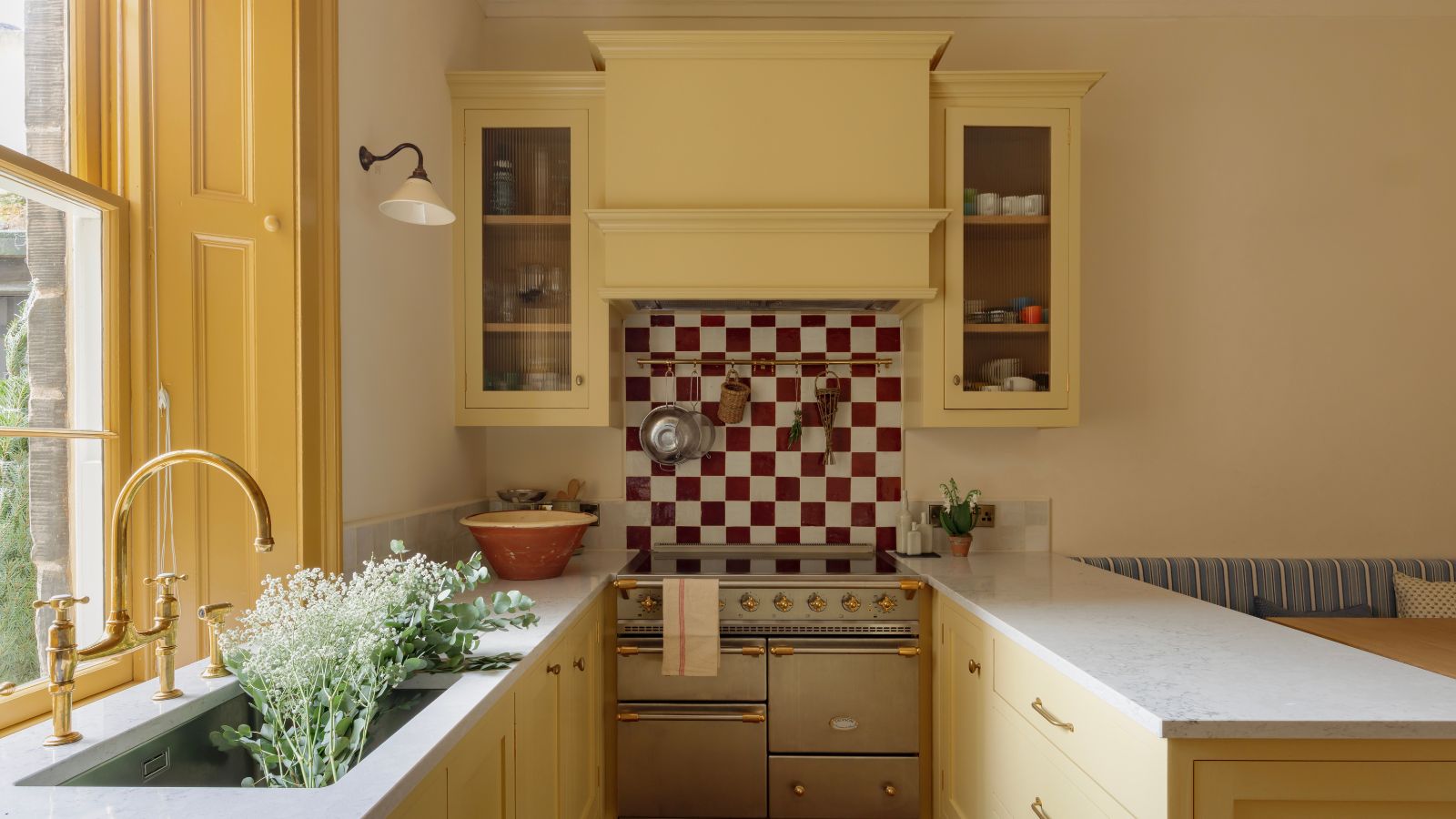Horseshoe layout kitchen with yellow Shaker cabinets, brass hardware, red and white checkerboard backsplash, farmhouse sink, and built-in banquette seating.
