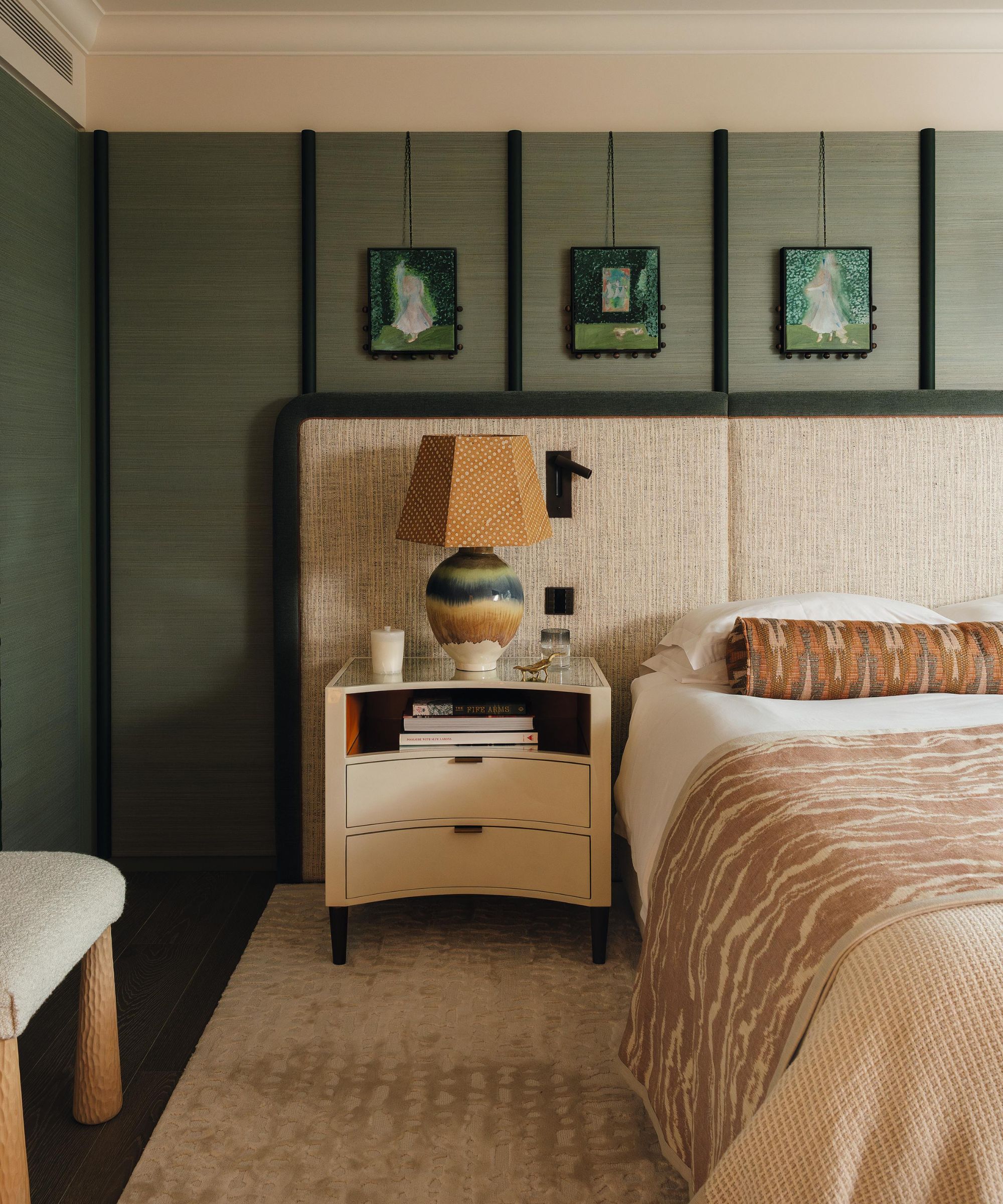 a green wallpapered bedroom with a large linen extended headboard, curved nightstand and unique wall art hanging from the picture rail