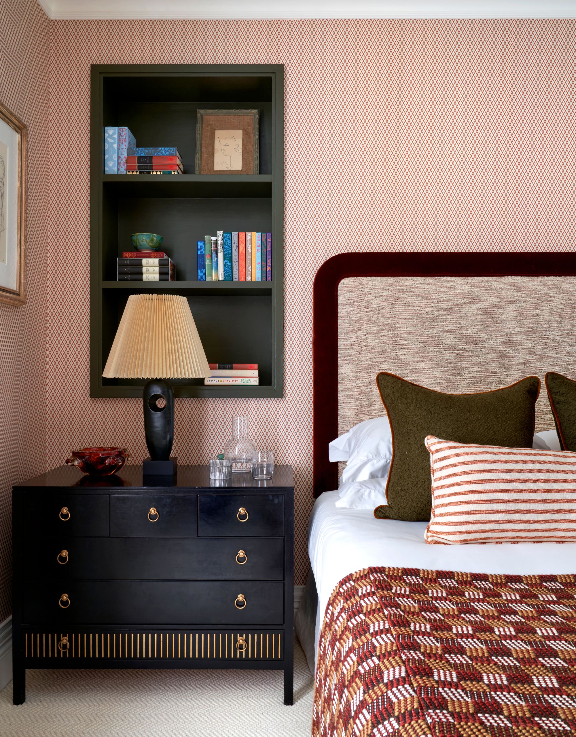 A modern bedroom with a subtle patterned headboard with a dark brown edge, a patterned throw and a dark blue chest of drawers with brass knobs below dark green-painted alcove shelving