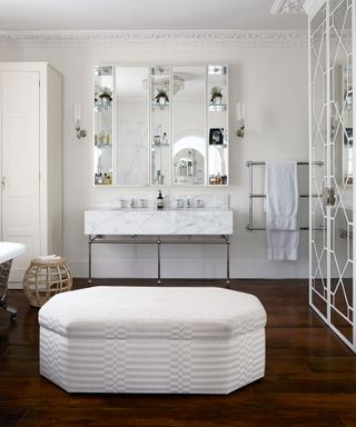 Bathroom with wooden floor, central white and grey ottoman, double marbled hand basin, elaborate cornice and chandelier.