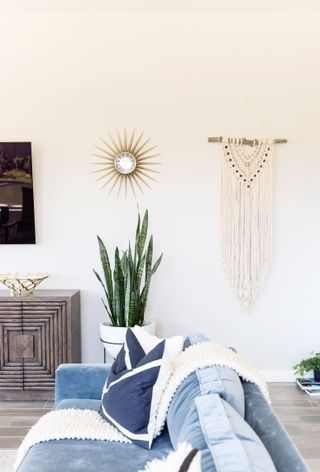white living room with blue couch, macrame on the wall, plant, starburst mirror, white textured throw, wooden carved side table