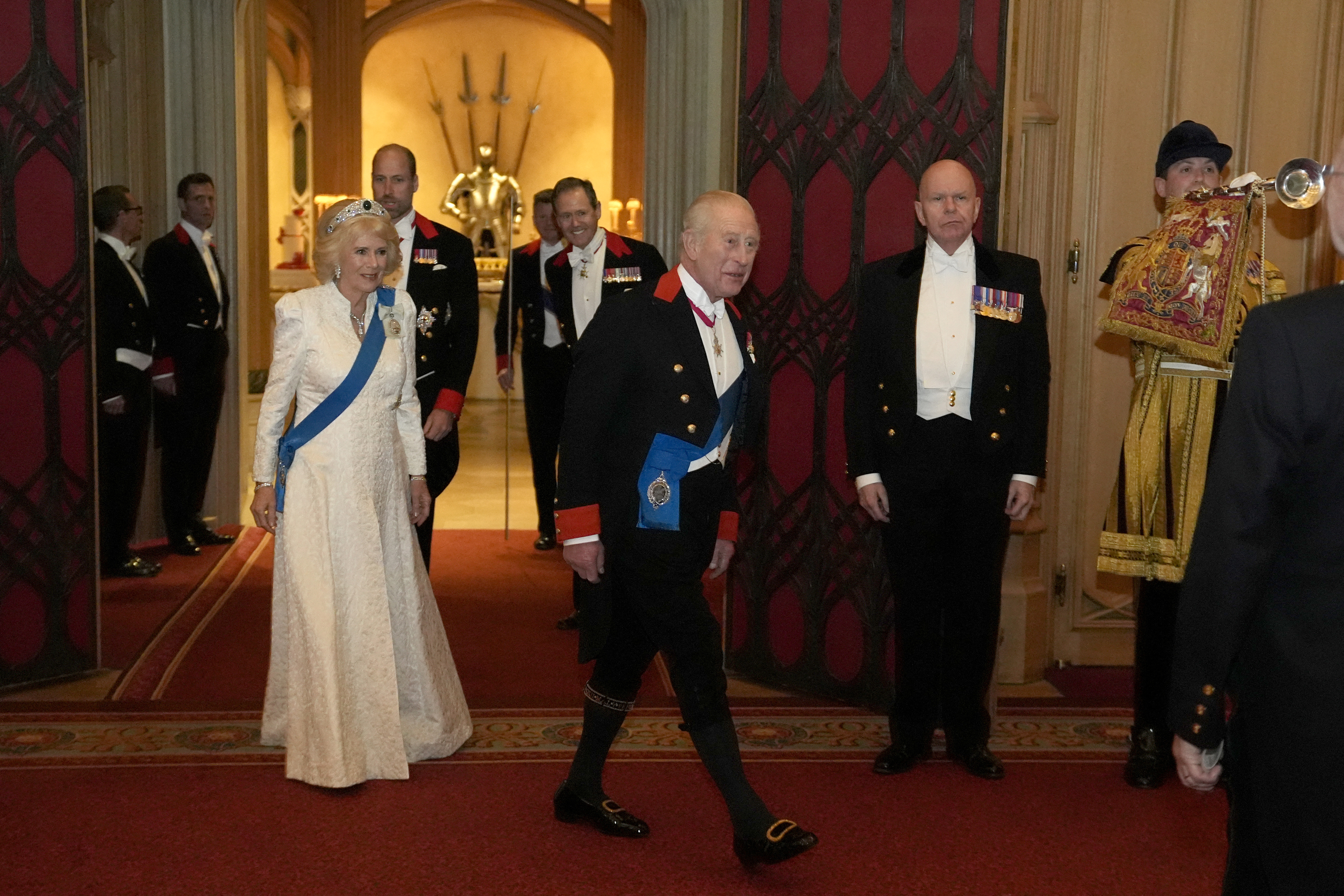 Queen Camilla wearing a white gown walking behind King Charles