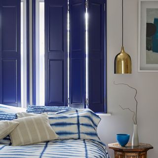 a bedroom with blue solid sghutters on the window and a bed in front covered in an ikat print blue and white bedlinen