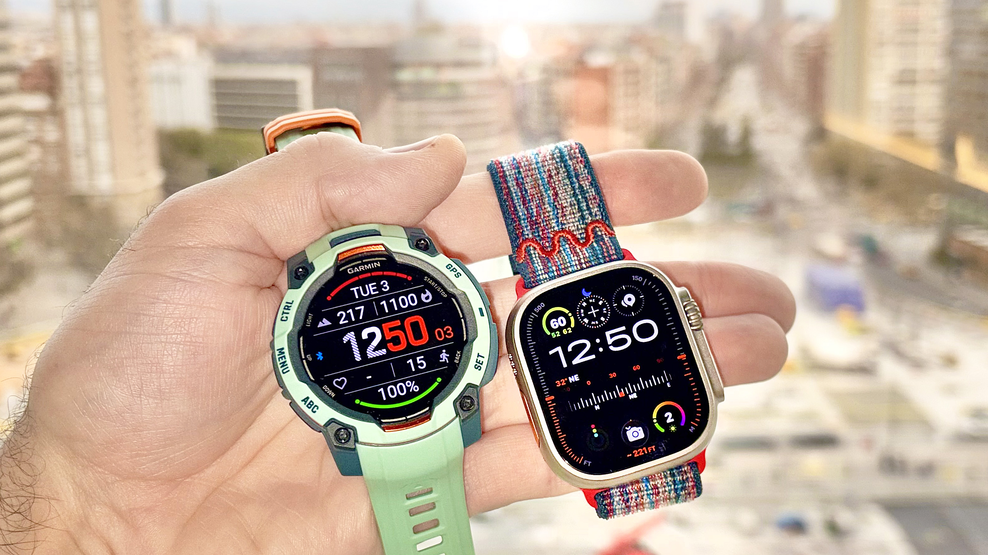Close-up of the Garmin Instinct 3 next to the Apple Watch Ultra 3 with the Barcelona skyline in the distance