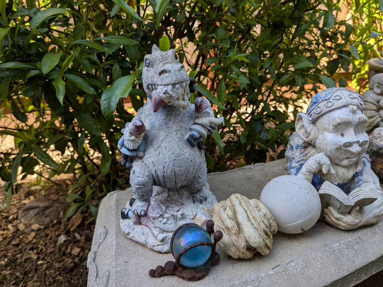 A Google Pixel 9a camera sample showing a dinosaur eating tiny gnomes next to a larger gnome reading a book.