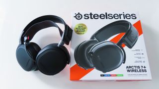 Ett par svarta SteelSeries Arctis 7+ ligger bredvid sin tillhörande förpackning på ett vitt bord.