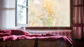 The image shows a wooden bedroom window open wide, with a bed with a red blanket on it below