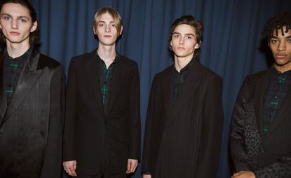 Four male models wearing looks from Paul Smith's collection. All four models are wearing black suit jackets in different styles and green, red, blue and black plaid style shirts